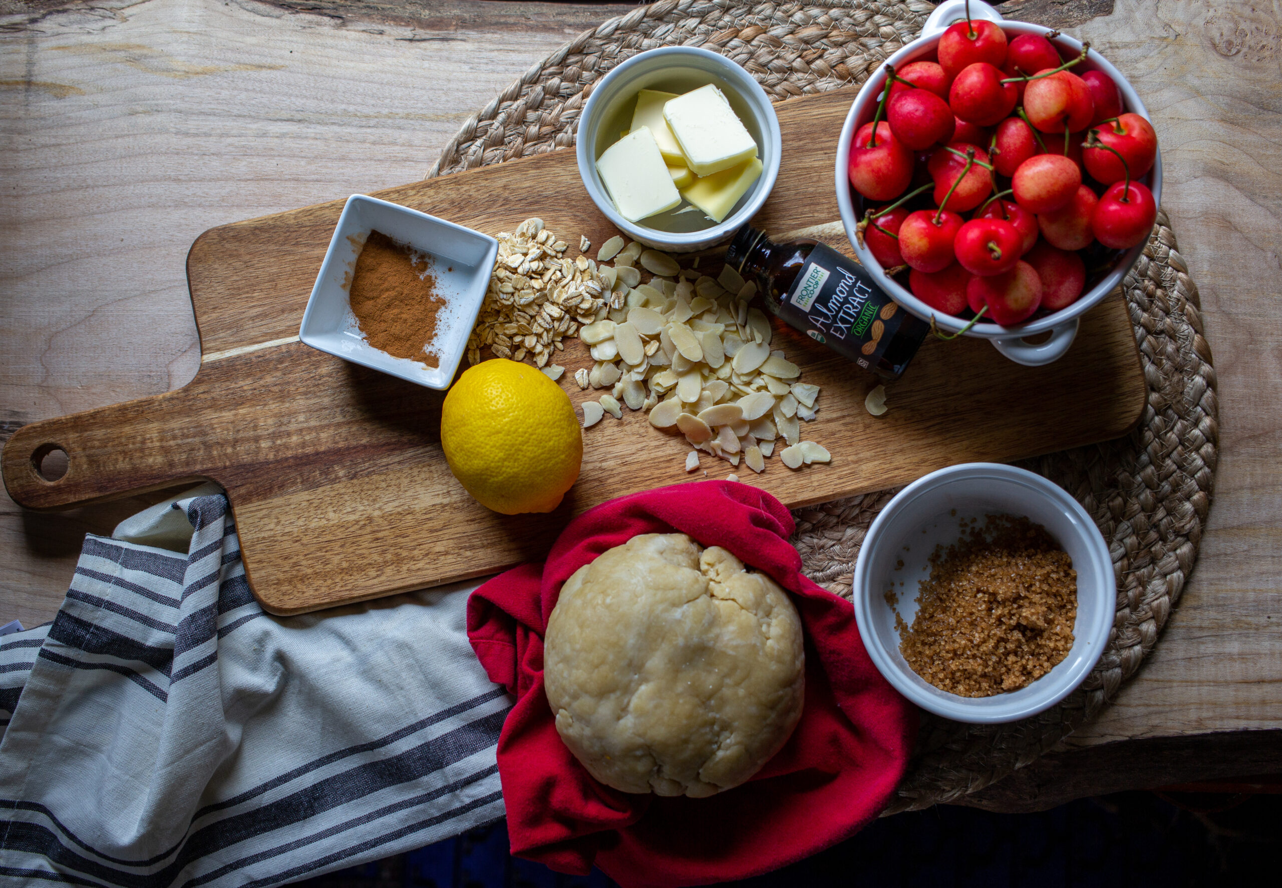 An assortment of ingredients including bowls of cherries, sugar, butter, lemon, a dough ball, and oats scattered on a wood board.