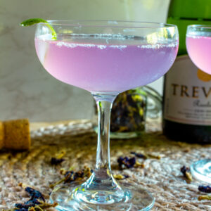 Purple cocktail in a coupe glass with a bottle of sparkling wine in the background.