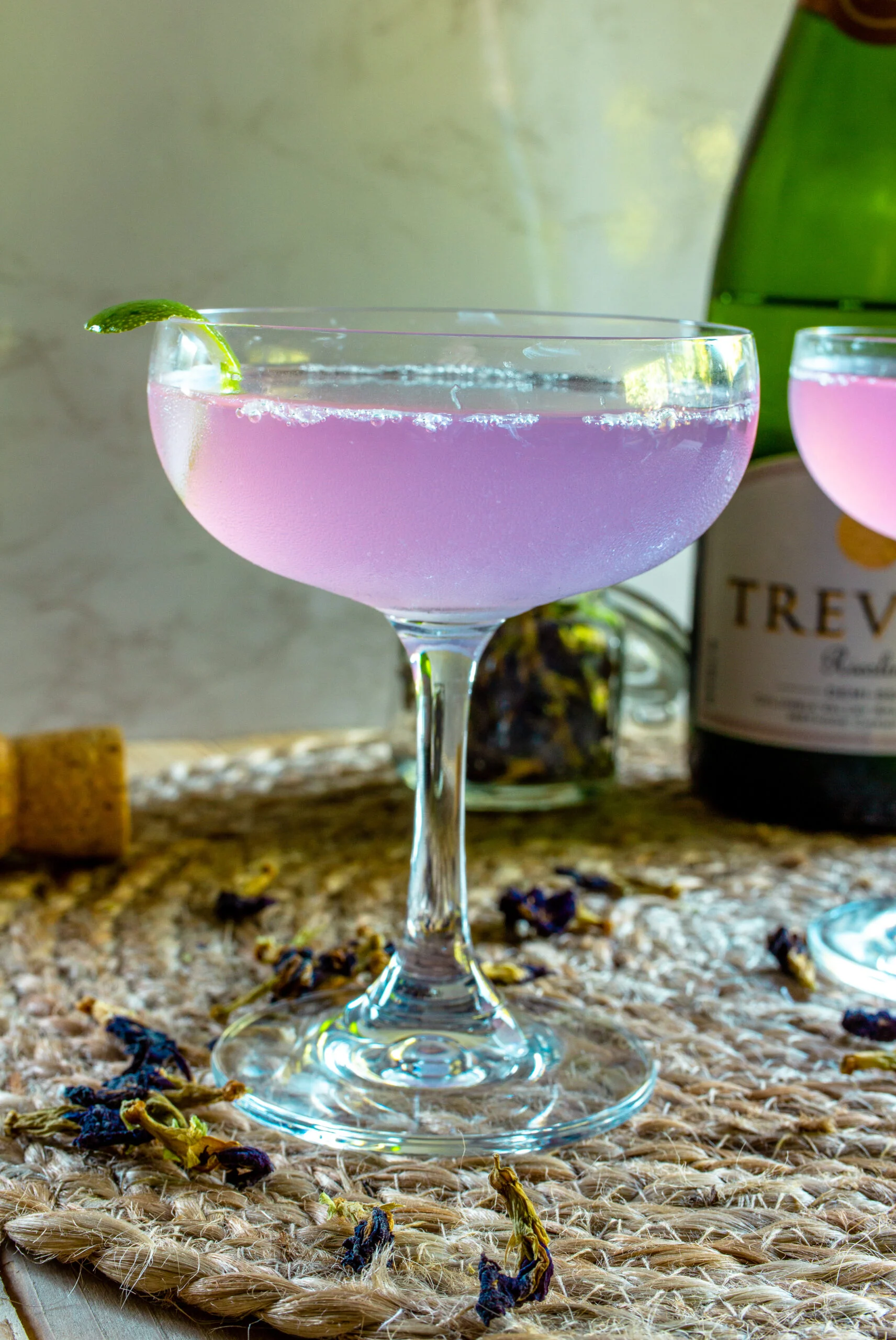 Purple cocktail in a coupe glass with a bottle of sparkling wine in the background.