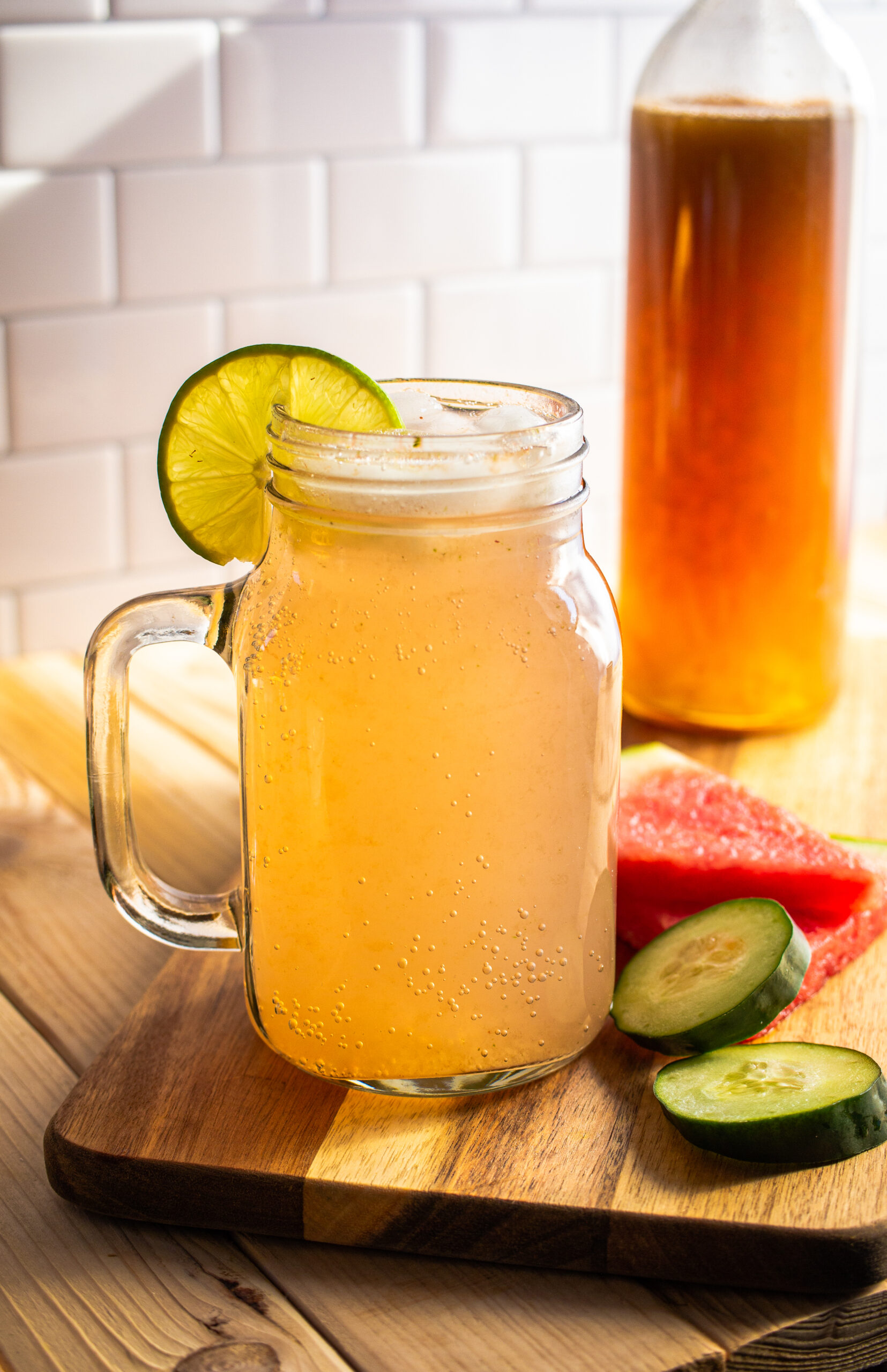 Cocktail in a mason jar on a board next to cucumbers and watermelon slices.