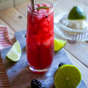 Cocktail in a tall glass with lime wedges and blackberries next to the glass.