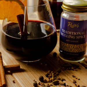 A glass of mulled wine with an orange wedge and cinnamon stick in the glass. A jar of mulling spices is next to it and has spilled the spices on the countertop.