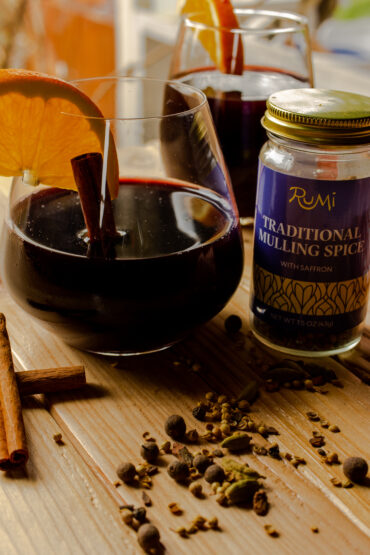 A glass of mulled wine with an orange wedge and cinnamon stick in the glass. A jar of mulling spices is next to it and has spilled the spices on the countertop.