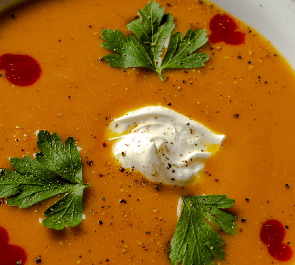Close up of carrot curry soup garnished with black pepper, sour cream, fresh parsley, and hot sauce.