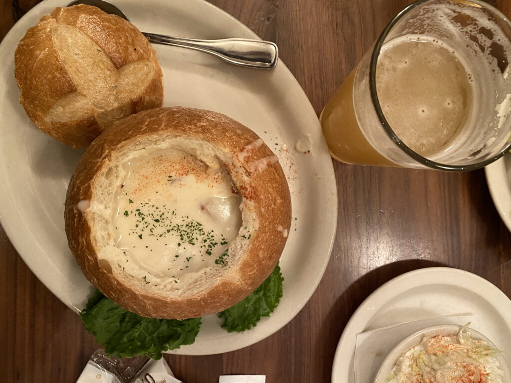 A soup and bread bowl on a white plate next to a beer.