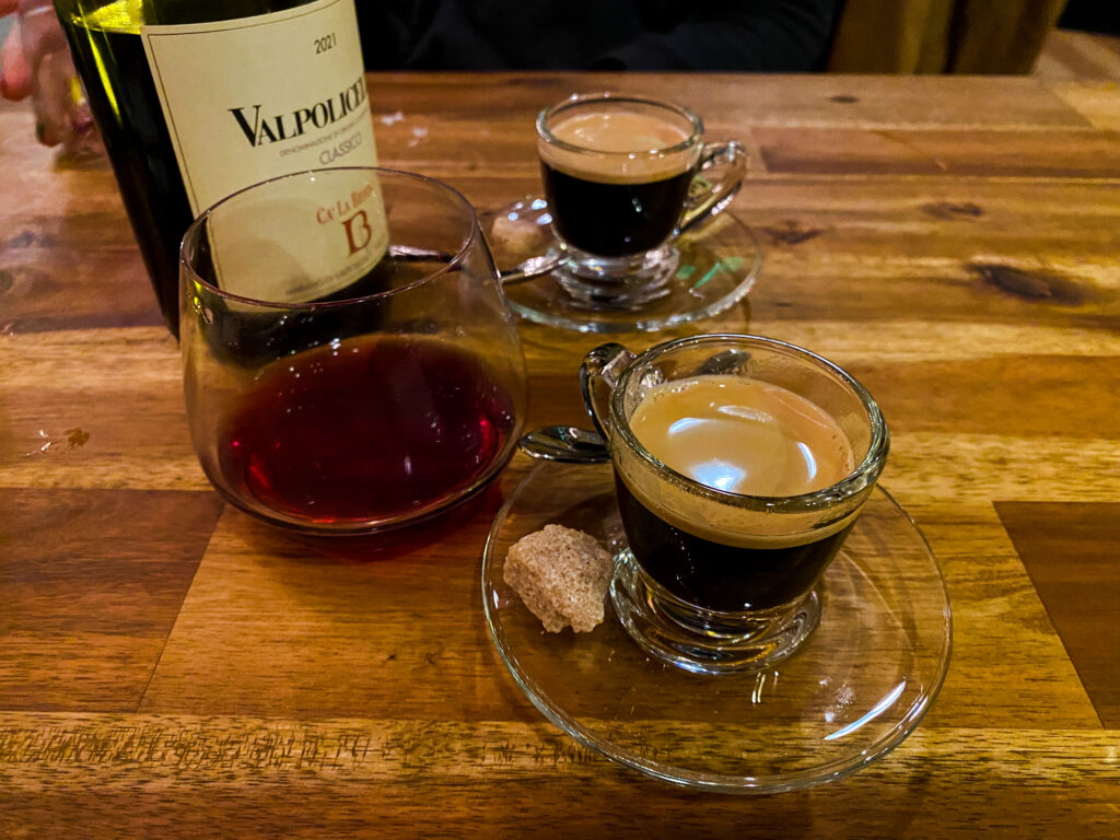 Two espressos on saucers next to a bottle of red wine and a glass of red wine, on a wood table.