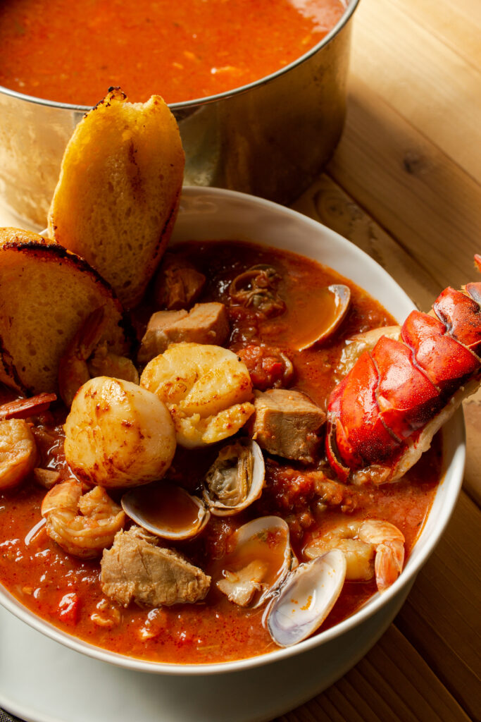 Cioppino in a white bowl with two wedges of toast in the bowl and the rest of the stew in the background.