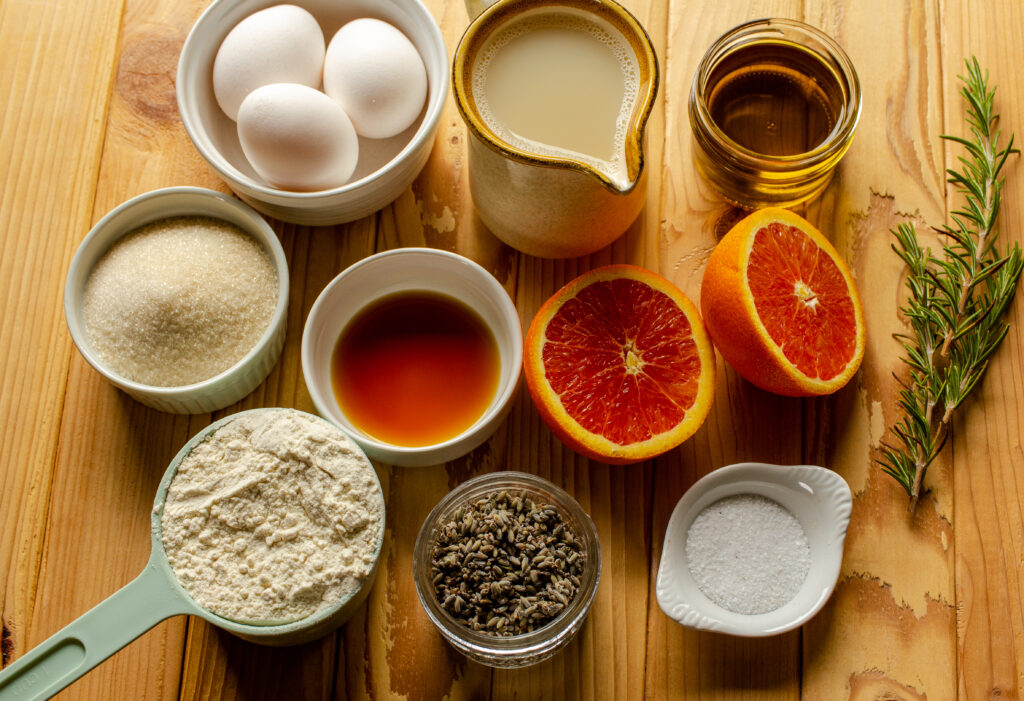 An assortment of ingredients for orange olive oil cake including oranges, vanilla, flour, lavender, salt, sugar, olive oil, and milk.