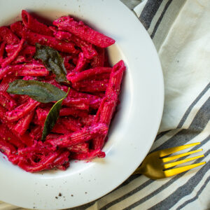Beet pasta with fried sage leaves in a white bowl with a fork next to the bowl.