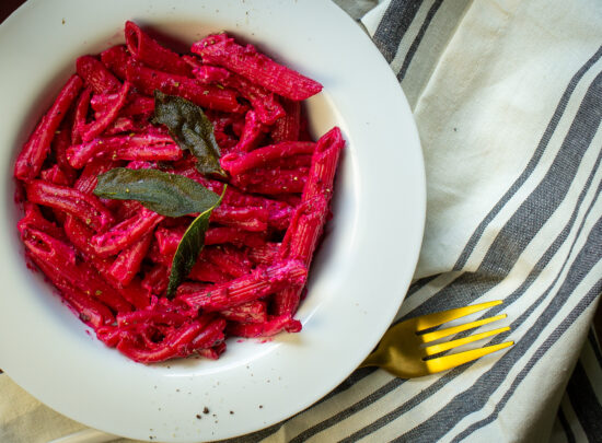 Beet pasta with fried sage leaves in a white bowl with a fork next to the bowl.