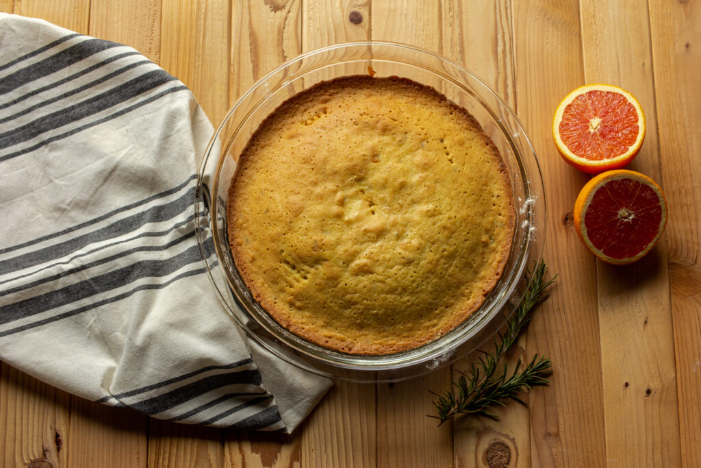 Orange olive oil cake in a clear cake pan surrounded by rosemary sprigs, a sliced orange, and a kitchen towel.