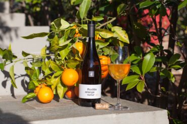 A bottle of wine next to an orange tree with a glass of orange wine.