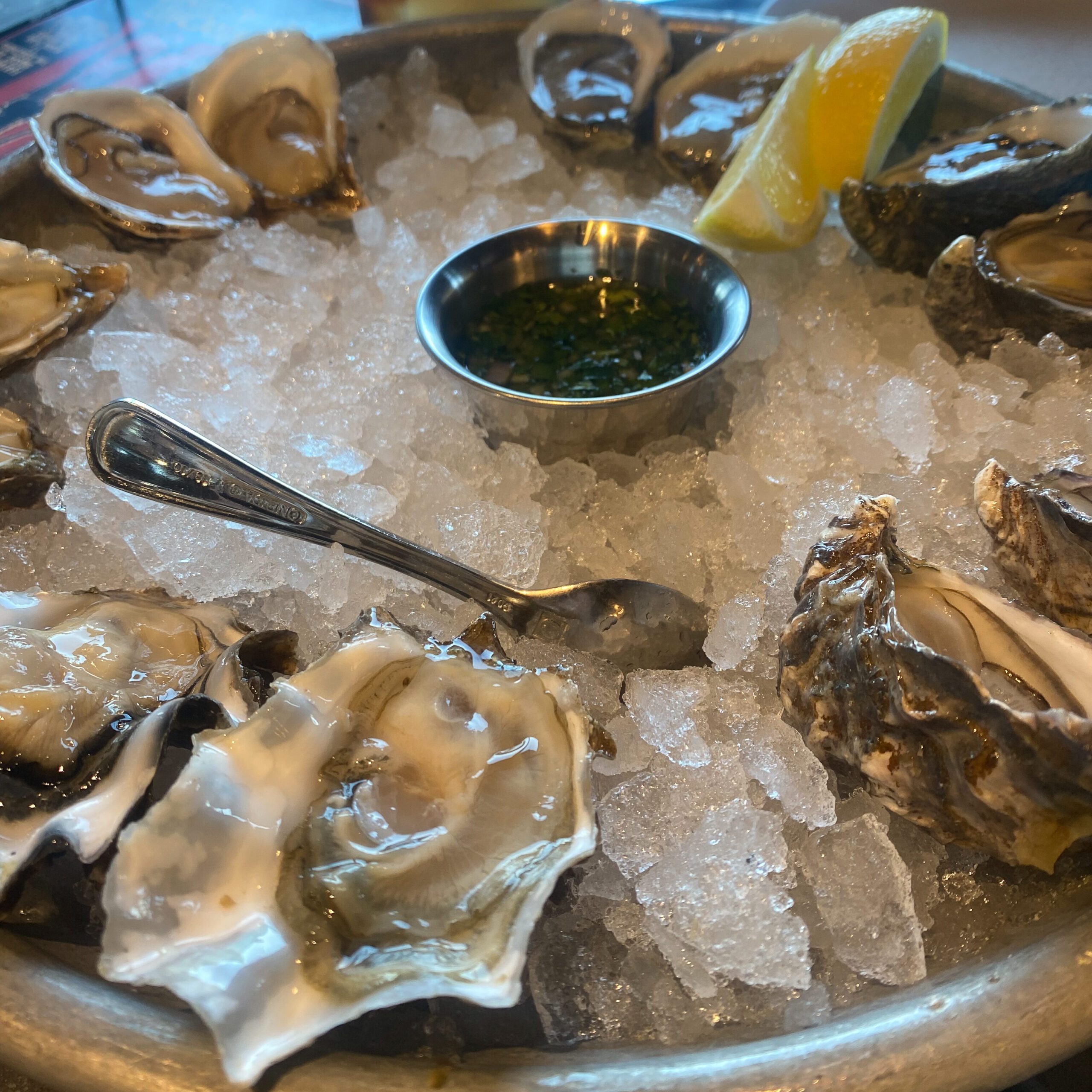A tray of oysters on a bed of ice with a spoon, lemon wedges, and a serving dish of mignonette in the bed of ice.