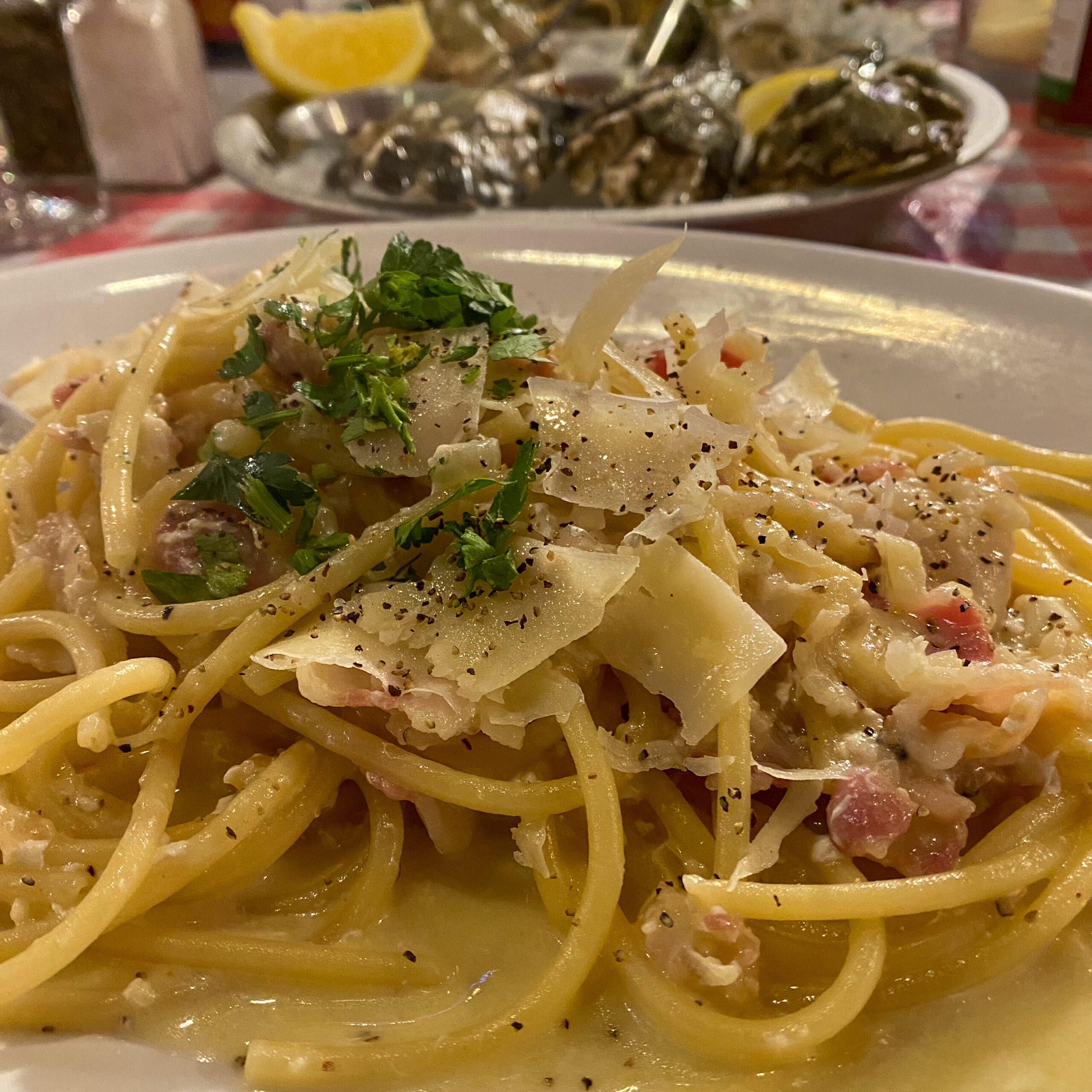 A serving of pasta carbonara garnished with black pepper and fresh herbs. In the background is a plate of oysters.