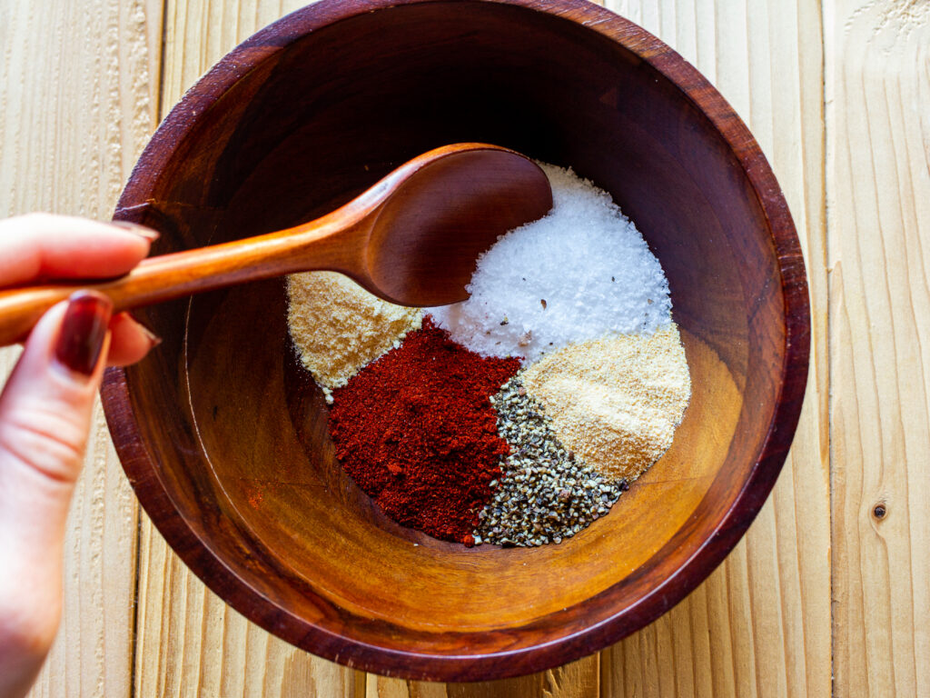 A wooden bowl filled with 5 different spices that have not yet been mixed together. A hand is holding a spoon about to mix the spices together.