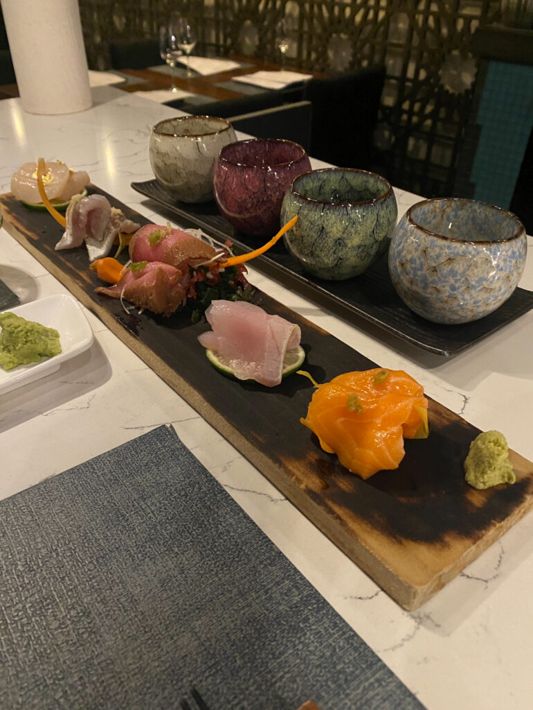 A burnt wood tray of sashimi next to a flight of multi-colored glasses.