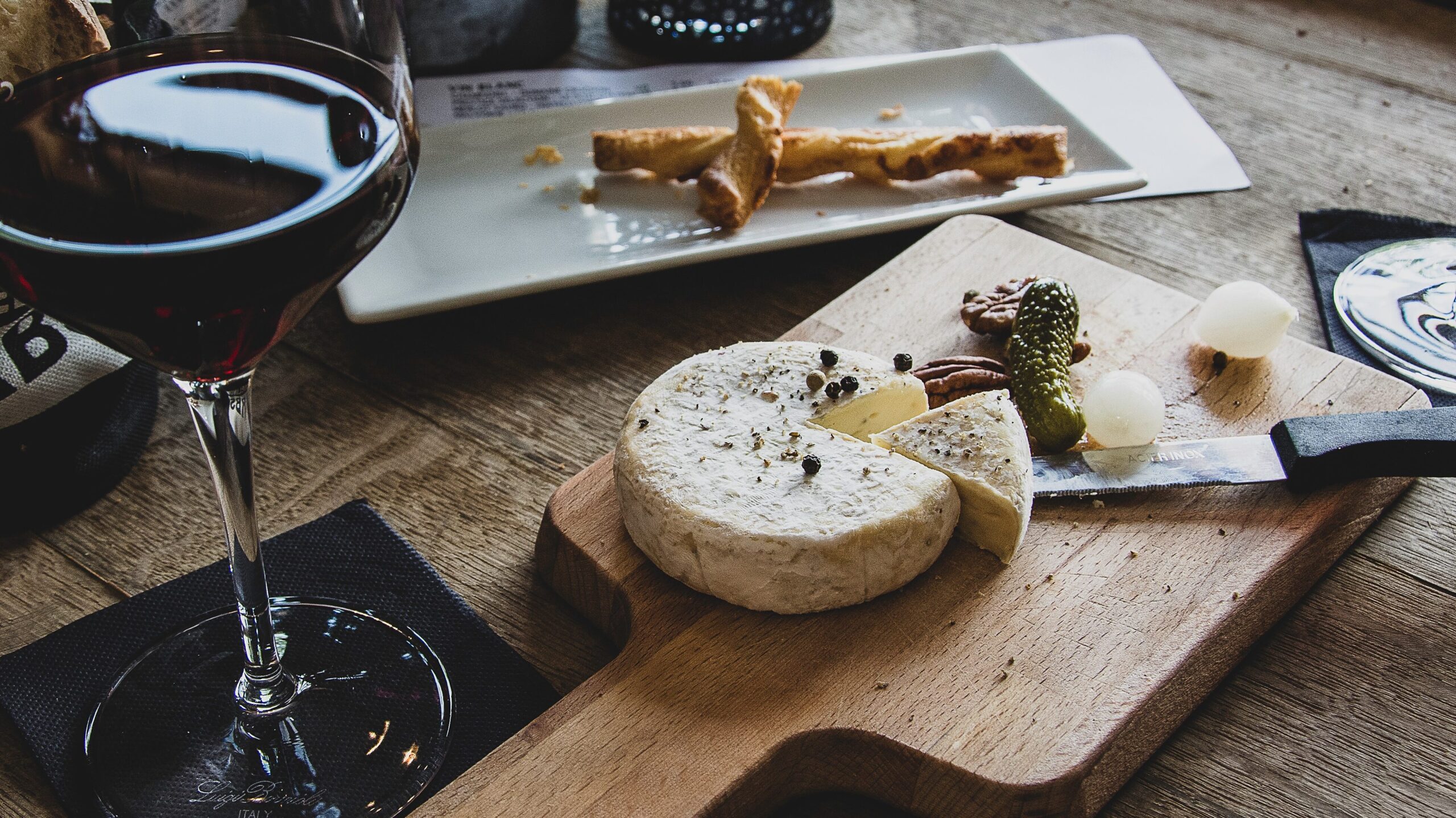 A charcuterie board with a cheese wheel, a knife, and a pickle, next to a glass of red wine and an empty plate.