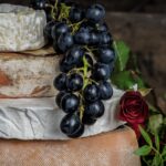 Four wheels of cheese stacked on each other with a bunch of grapes atop and roses on the side.