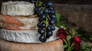 Four wheels of cheese stacked on each other with a bunch of grapes atop and roses on the side.