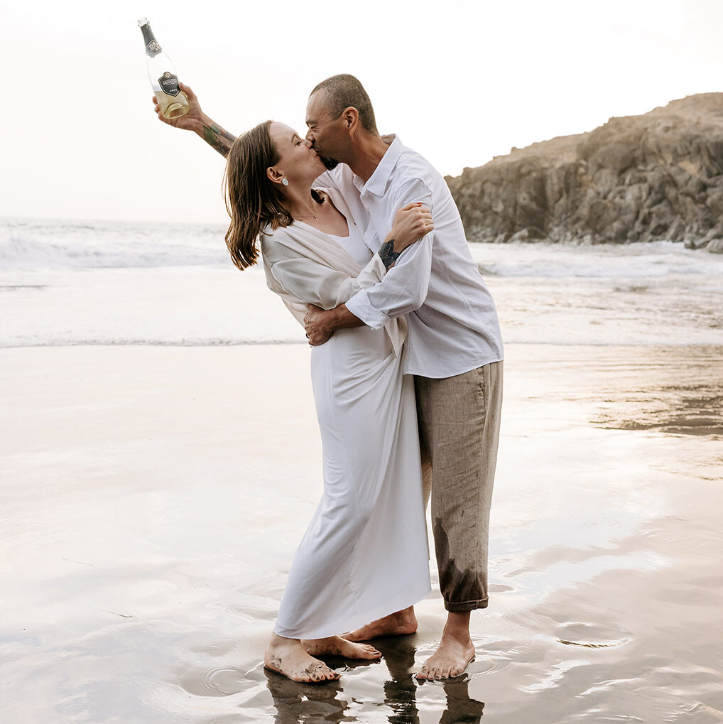 A husband and wife kissing on the beach and holding a bottle of sparkling wine.