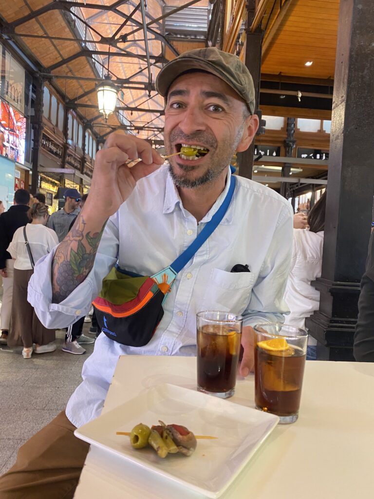 A man eating an olive in a market with two glasses of vermouth.